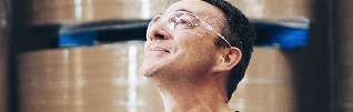 Factory worker wearing safety glasses and earplugs smiles, with large paper rolls in the background.