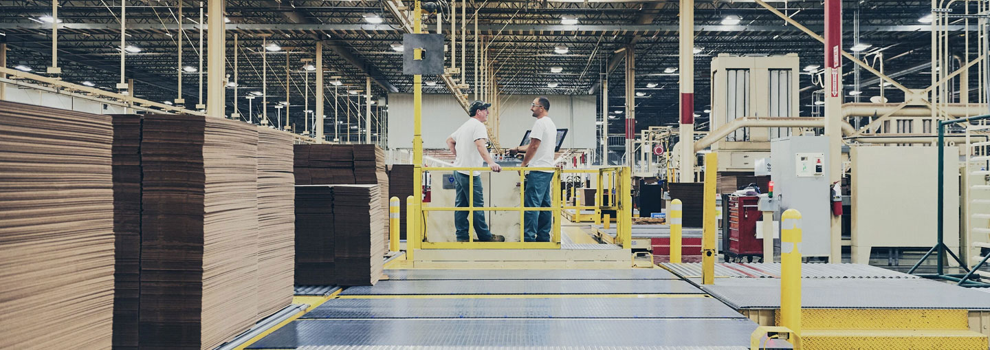 Cardboard manufacturing plant with employees in production and stacks of corrugated cardboard on an industrial line
